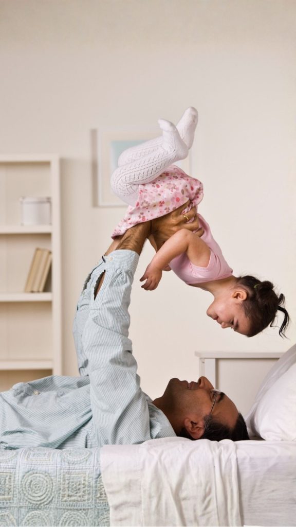 A father playfully lifting his young daughter in the air while lying on a bed inside their bright Orlando home.