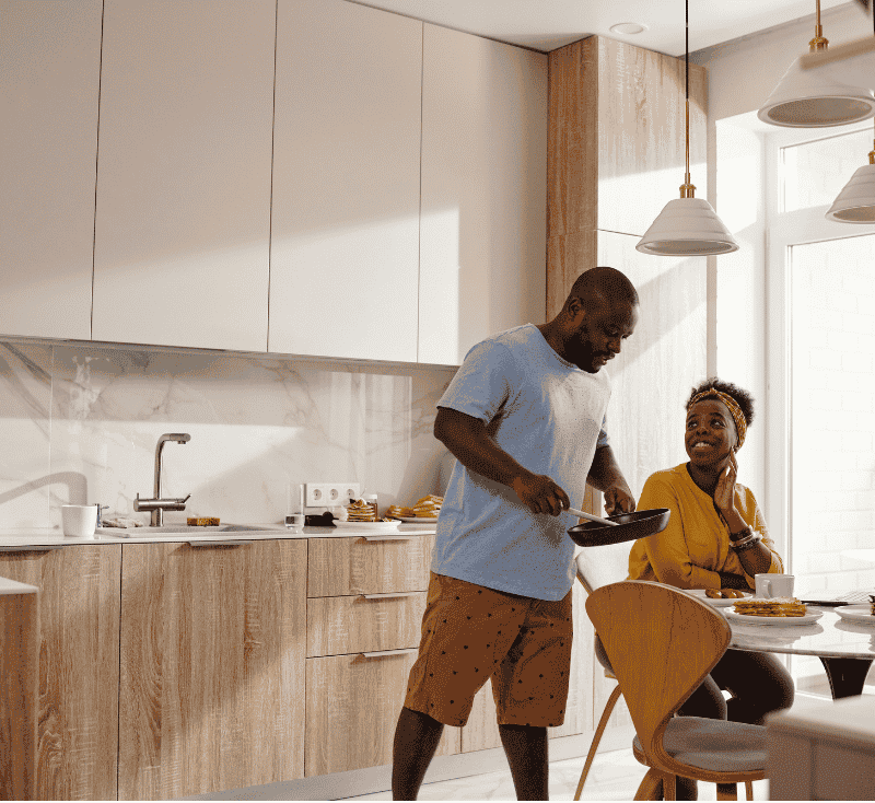 Man cooking pancakes in a bright kitchen while a smiling woman sits at the table with a laptop