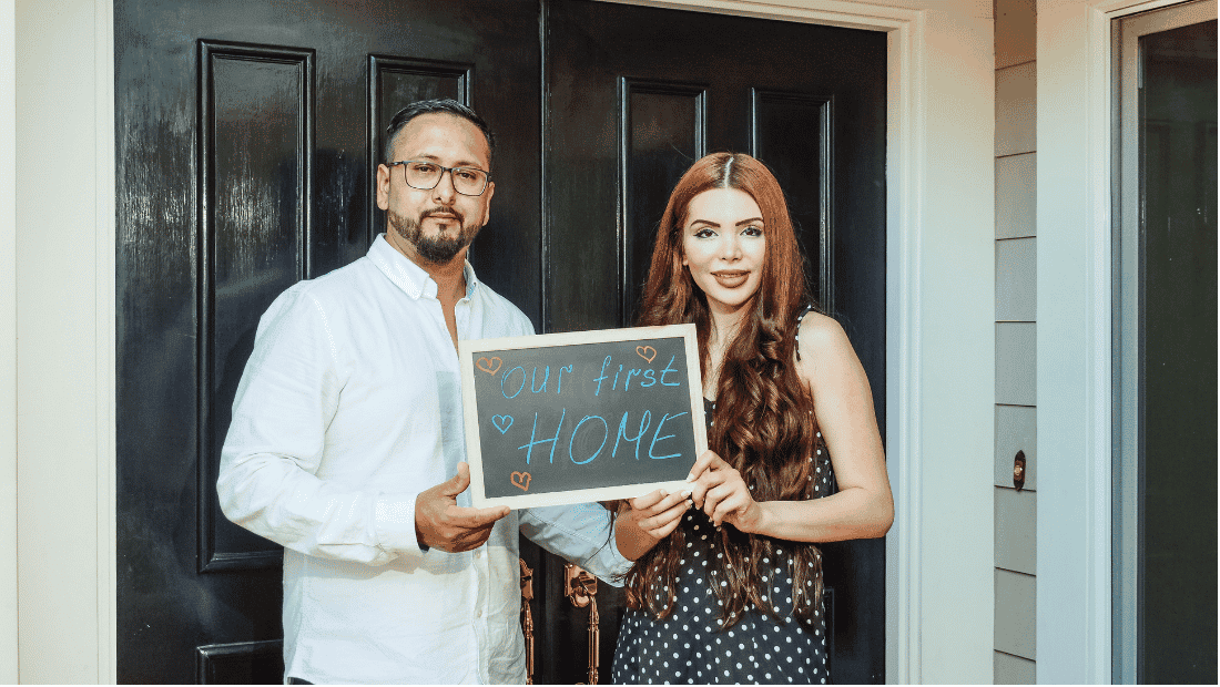 Couple standing at a black front door holding a small chalkboard that reads "Our first home"