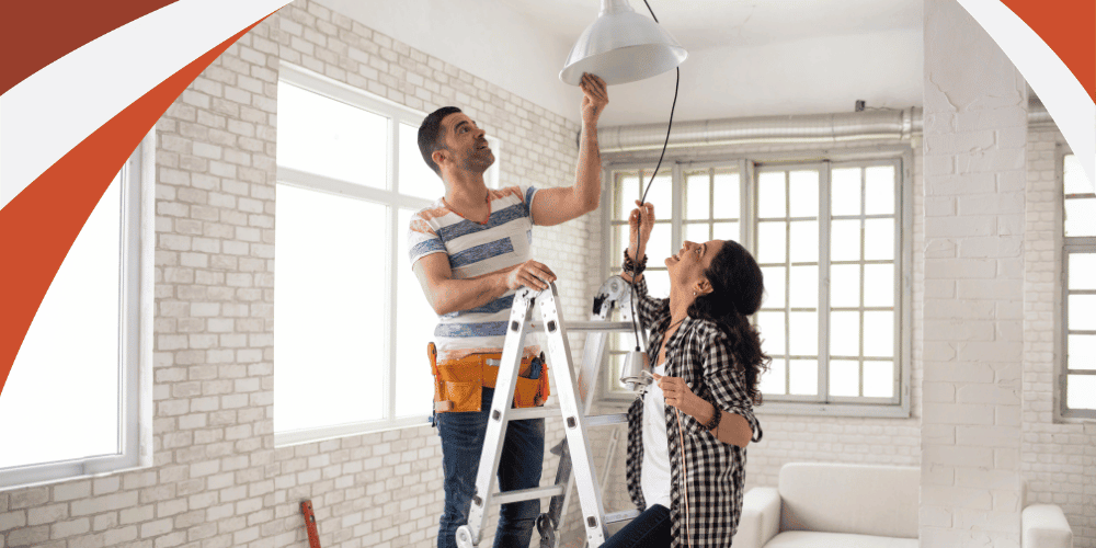 Man on a stepladder holding a pendant light while a woman beside him guides the cord in a room with white brick walls