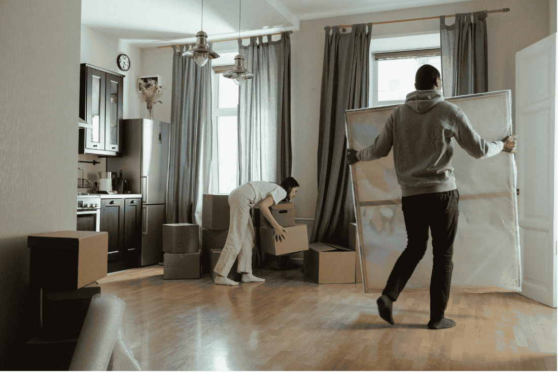 Woman lifting a cardboard moving box while a man carries a large wrapped canvas across a living room filled with boxes