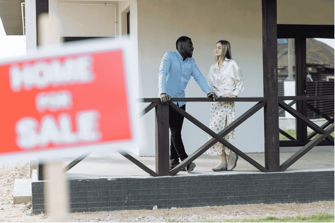 Couple talking on the porch of a white house, with a blurred red "Home for Sale" sign in the foreground