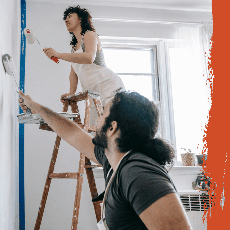 Woman on a wooden ladder and a man beside her painting a white wall with paint rollers