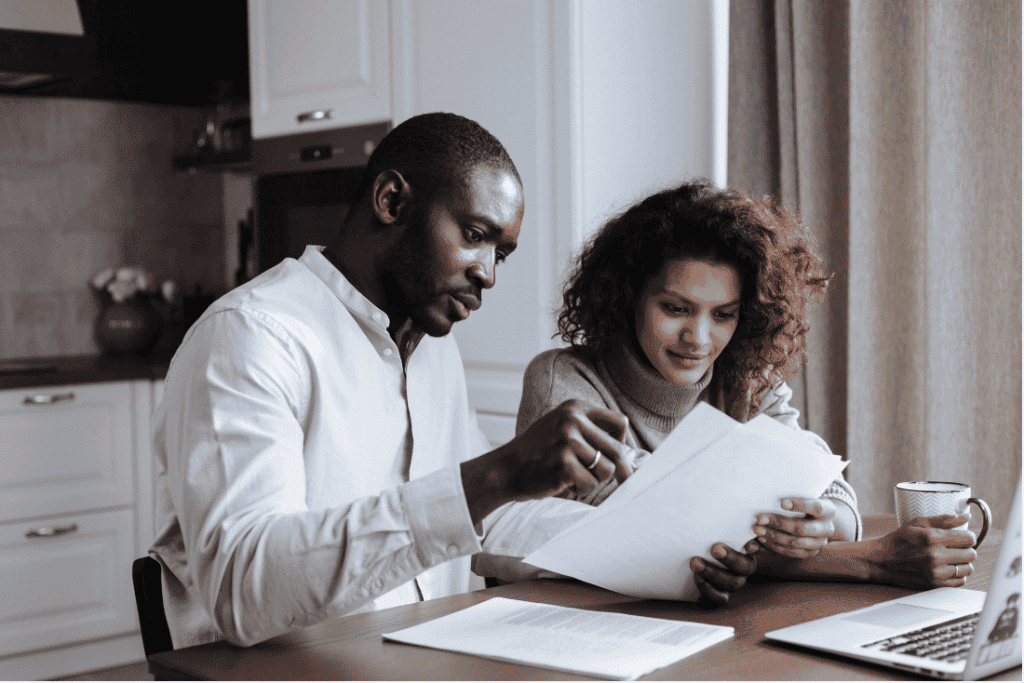 Man and woman sitting at a kitchen table reading a sheet of paper together, with more papers on the table in front of them