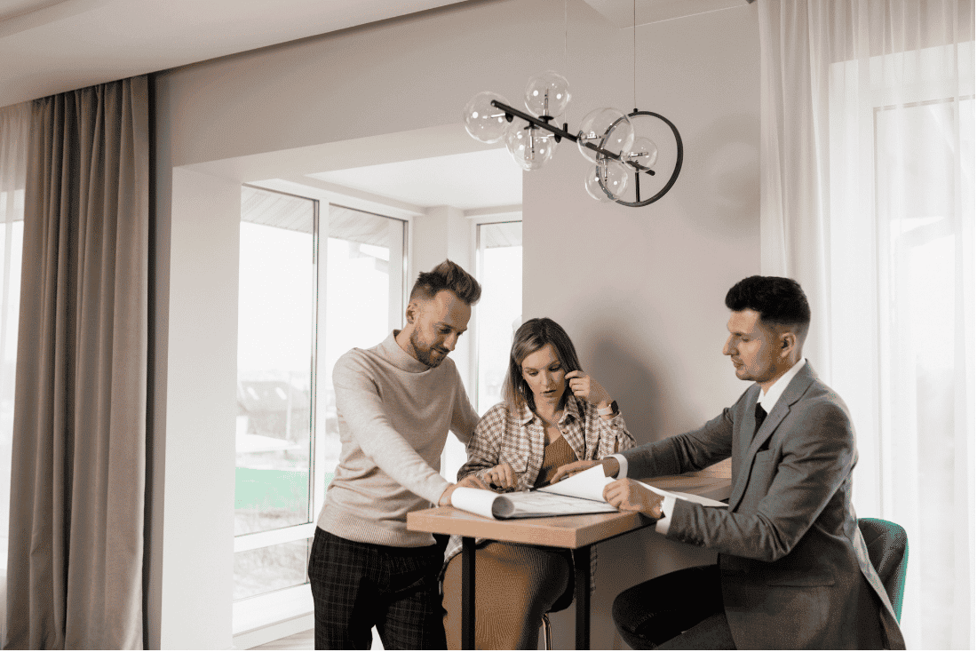 Couple looking over rolled plans at a high table with a man in a gray suit, in a bright room with large windows