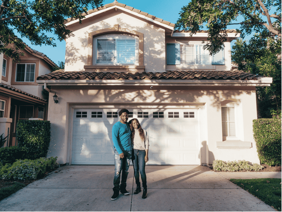 Smiling couple standing with their arms around each other in the driveway of a two-story stucco house with a tile roof