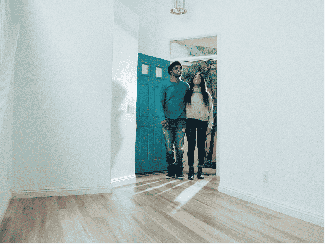 Smiling couple standing just inside the open teal front door of an empty house with white walls and light wood floors