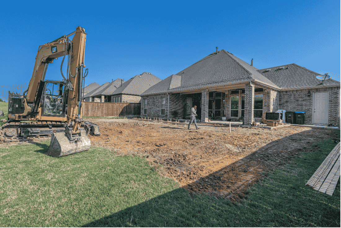 Excavator beside a cleared dirt area in the backyard of a single-story brick house, with a worker walking near the covered patio