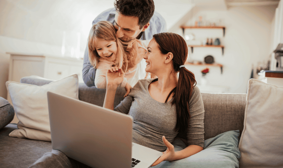 Family laughing together on the living room couch while the mother works on a laptop