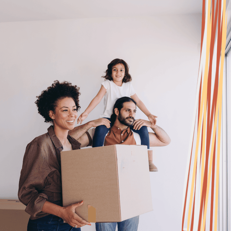Smiling woman carrying a cardboard moving box beside a man with a young girl on his shoulders in a bright room