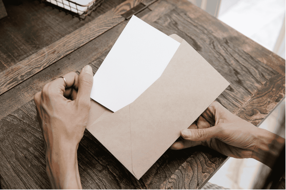 Hands pulling a blank white card out of a brown paper envelope over a wooden desk