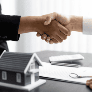 Two people shaking hands over a desk with a model house, house keys and paperwork