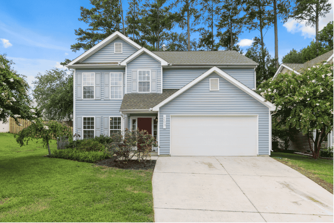 Light blue two-story house with a red front door, a white garage door and a landscaped front yard, with tall pine trees behind it