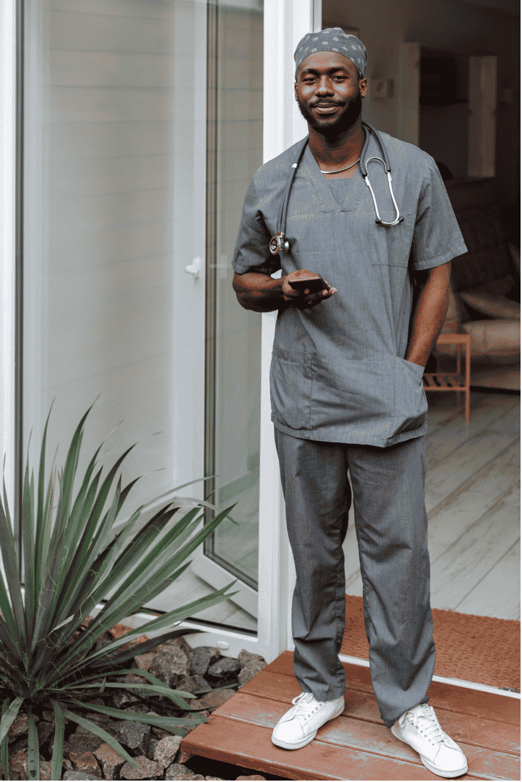 Smiling man in gray scrubs and a scrub cap, with a stethoscope around his neck, standing in the open doorway of a home