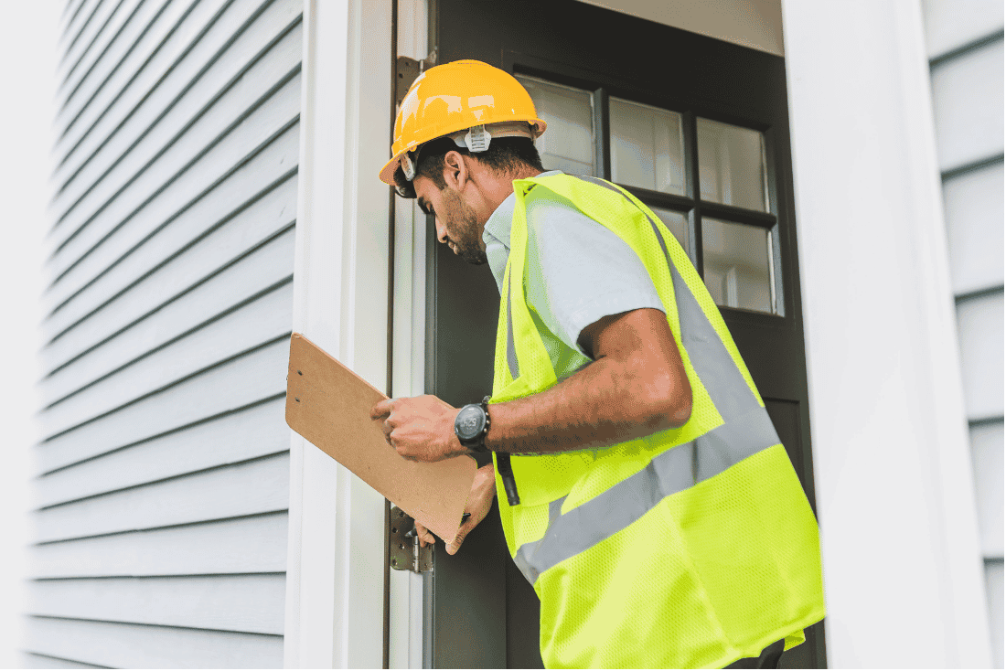 Man in a yellow hard hat and safety vest holding a clipboard while checking the front door frame of a house with gray siding