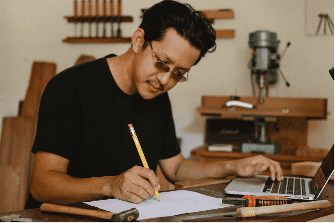 Man in a black T-shirt and tinted glasses writing on paper with a pencil at a workbench in a woodworking shop, with one hand on a laptop