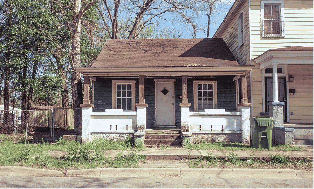 Small older bungalow with dark green siding, a worn front porch and weeds growing along the sidewalk