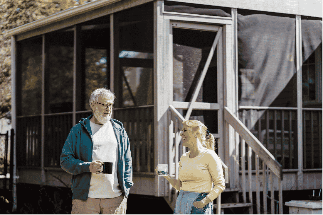 Older man and woman holding coffee mugs and talking outside a screened porch with wooden steps