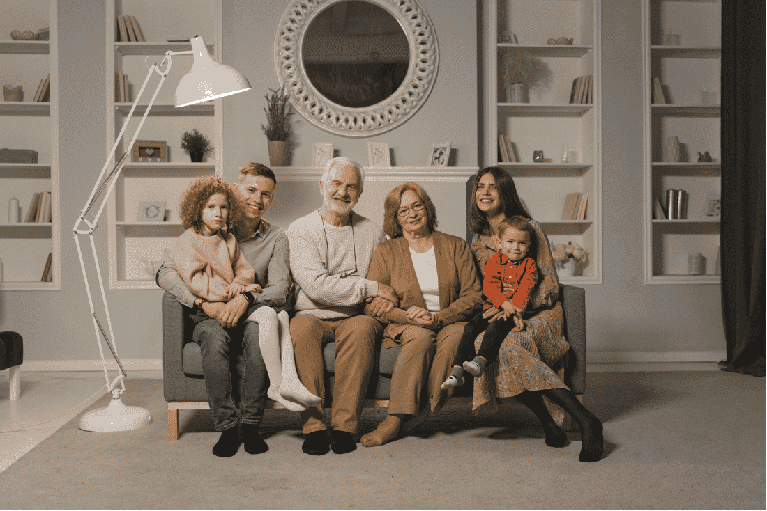 Older couple sitting on a gray sofa between a younger man and woman, each holding a small child, in a living room with white bookshelves
