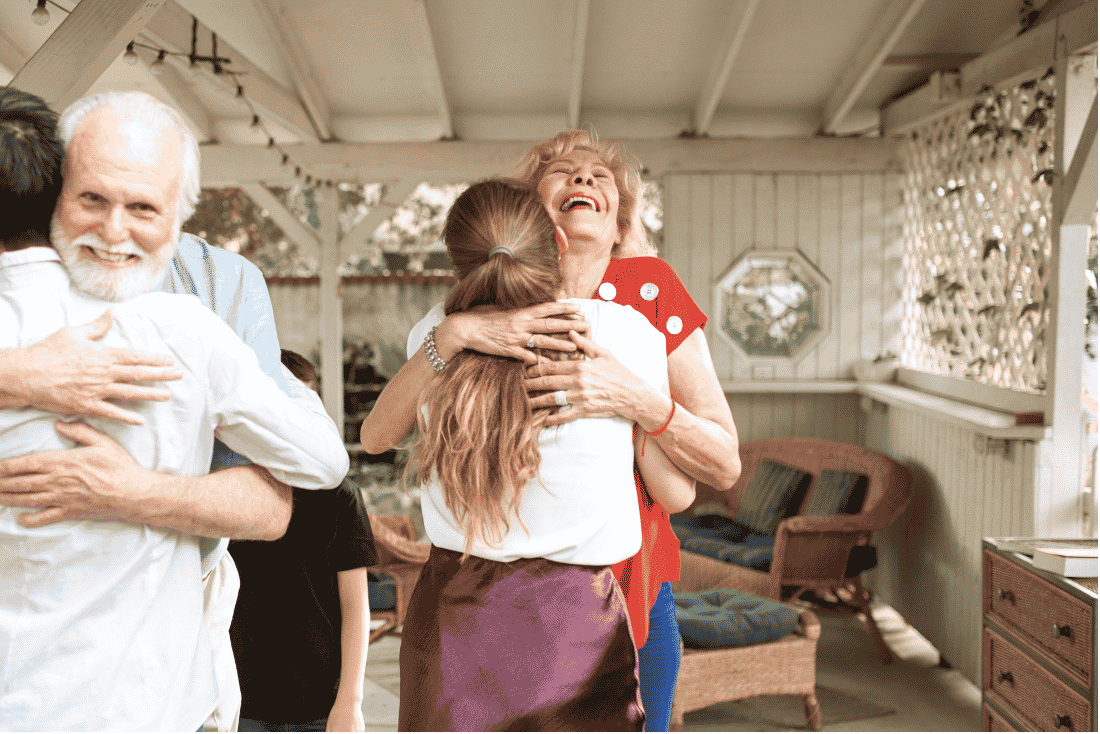 Older man with a white beard and a laughing older woman each hugging a younger person on a covered white porch