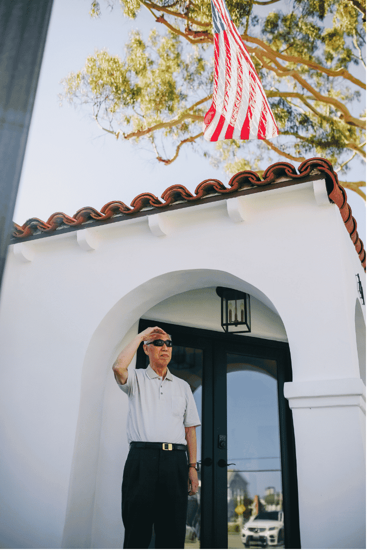 Older man in sunglasses with his hand raised to his brow outside a white stucco entry with a clay tile roof and an American flag flying above