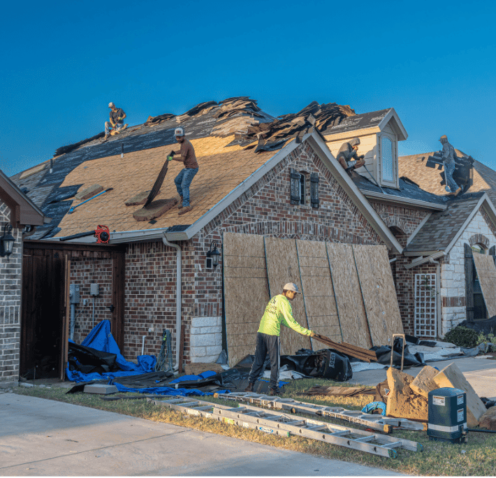 Roofing crew removing old shingles from the roof of a brick house under a clear blue sky