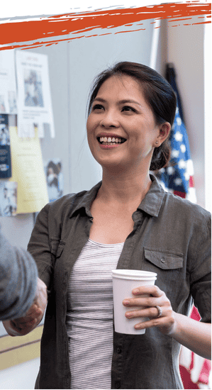 Smiling woman holding a coffee cup and shaking hands, with an American flag on the wall behind her