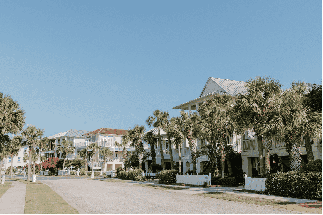 Residential street lined with palm trees and multi-story houses with porches, balconies and metal and tile roofs under a clear sky