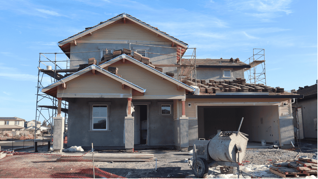 Two-story house under construction with scaffolding, unfinished stucco walls and tiles stacked on the roof