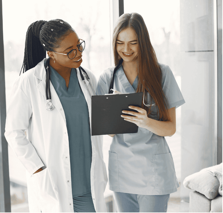Two smiling women with stethoscopes, one in a white coat and one in light blue scrubs, looking at a clipboard beside a bright window