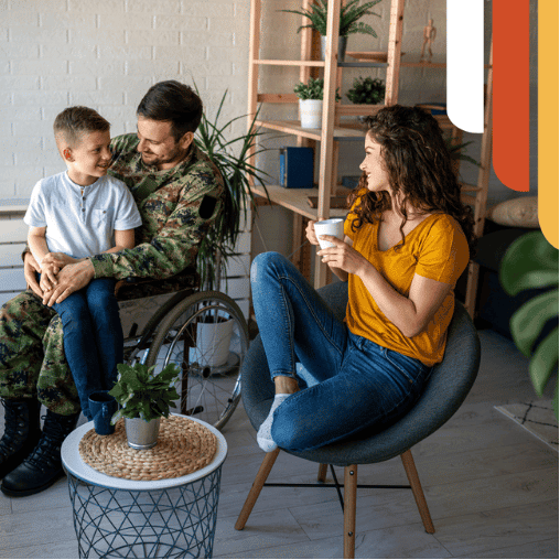 Service member in camouflage uniform in a wheelchair holding his son while his wife smiles with a mug at home