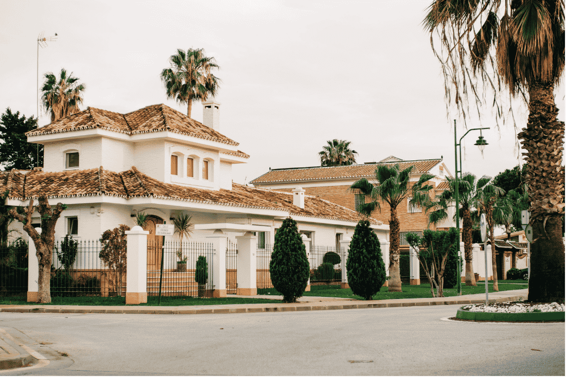 White two-story Mediterranean-style house with a clay tile roof behind a wrought-iron fence, with palm trees around it