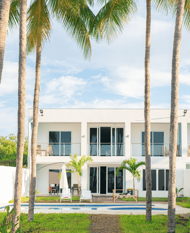 White two-story house with balconies and a swimming pool in front, framed by tall palm trees under a bright sky