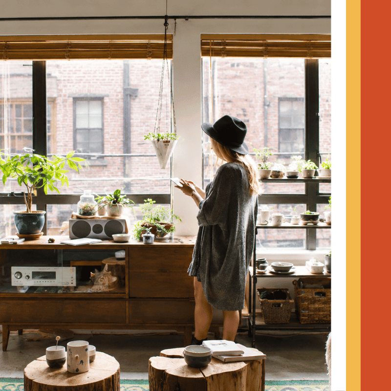 Woman in a hat looking at her phone by large windows in a sunny apartment filled with plants