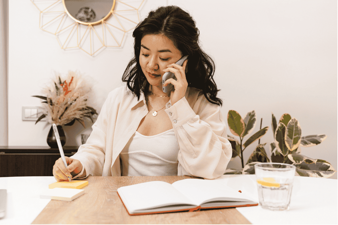 Woman talking on a phone while writing on a sticky note at a table, with an open notebook in front of her