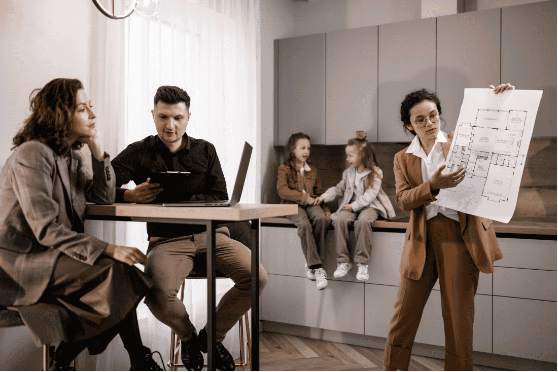 Woman holding up a floor plan for a couple seated at a table, with two children sitting on a kitchen counter behind them
