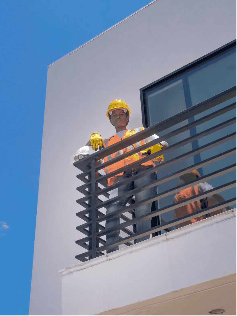 Worker in a yellow hard hat and orange safety vest standing on the balcony of a white stucco building against a blue sky