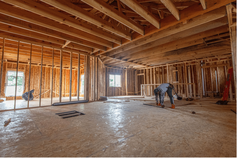 Worker bending over the subfloor inside a wood-framed house under construction, with exposed wall studs and ceiling joists